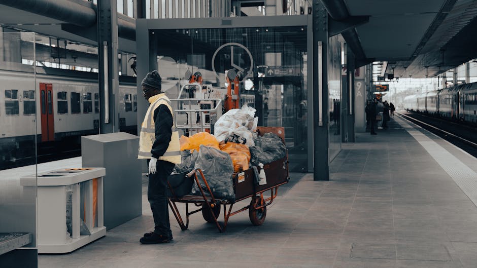 At a modern train station platform, a man dressed in dark clothing, wearing a beanie and a high-visibility vest, stands beside a large red hand truck loaded with various bags of rubbish, including bla