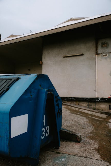 At a modern train station platform, a man dressed in dark clothing, wearing a beanie and a high-visibility vest, stands beside a large red hand truck loaded with various bags of rubbish, including bla